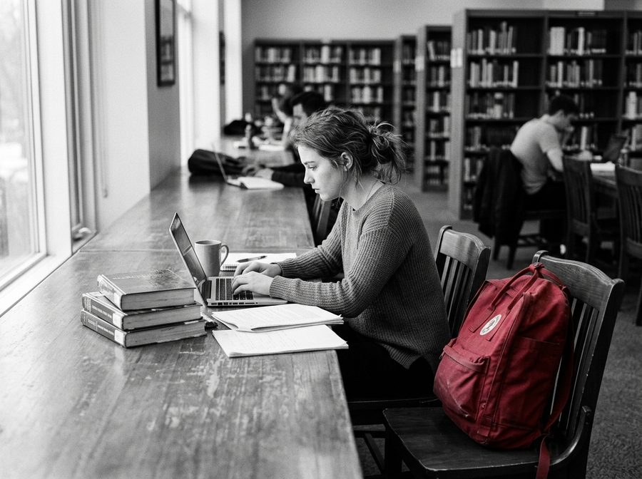 Une étudiante travaille à une table de bibliothèque avec un ordinateur portable, un sac à dos rouge sur la chaise à côté d’elle