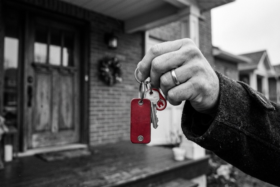Une main tient des clés de maison sur un porte-clés rouge, devant une maison de briques