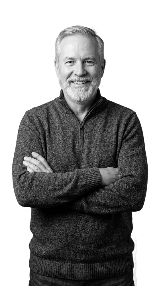 Un homme plus âgé et soulagé à la barbe grise, les bras croisés, photographié en noir et blanc sur un fond rouge érable.