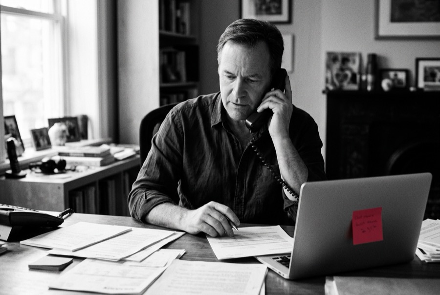 A man on the phone at a desk covered in bills and paperwork
